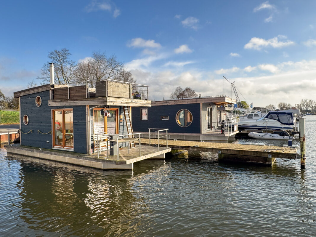 Zwei Hausboote, Holzbauweise, festliegend an einem Steg bei Sonnenschein. Liegende Motorboote im Hintergrund