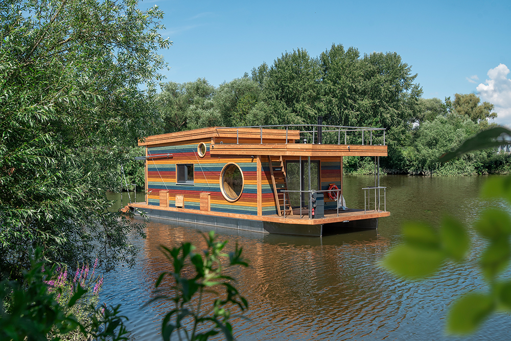 Modernes dezent bunt gestreiftes Hausboot frei schwimmend auf ruhigem Gewässer. Diagonalsicht auf Bug mit Steuerstand und Dachterrasse.
