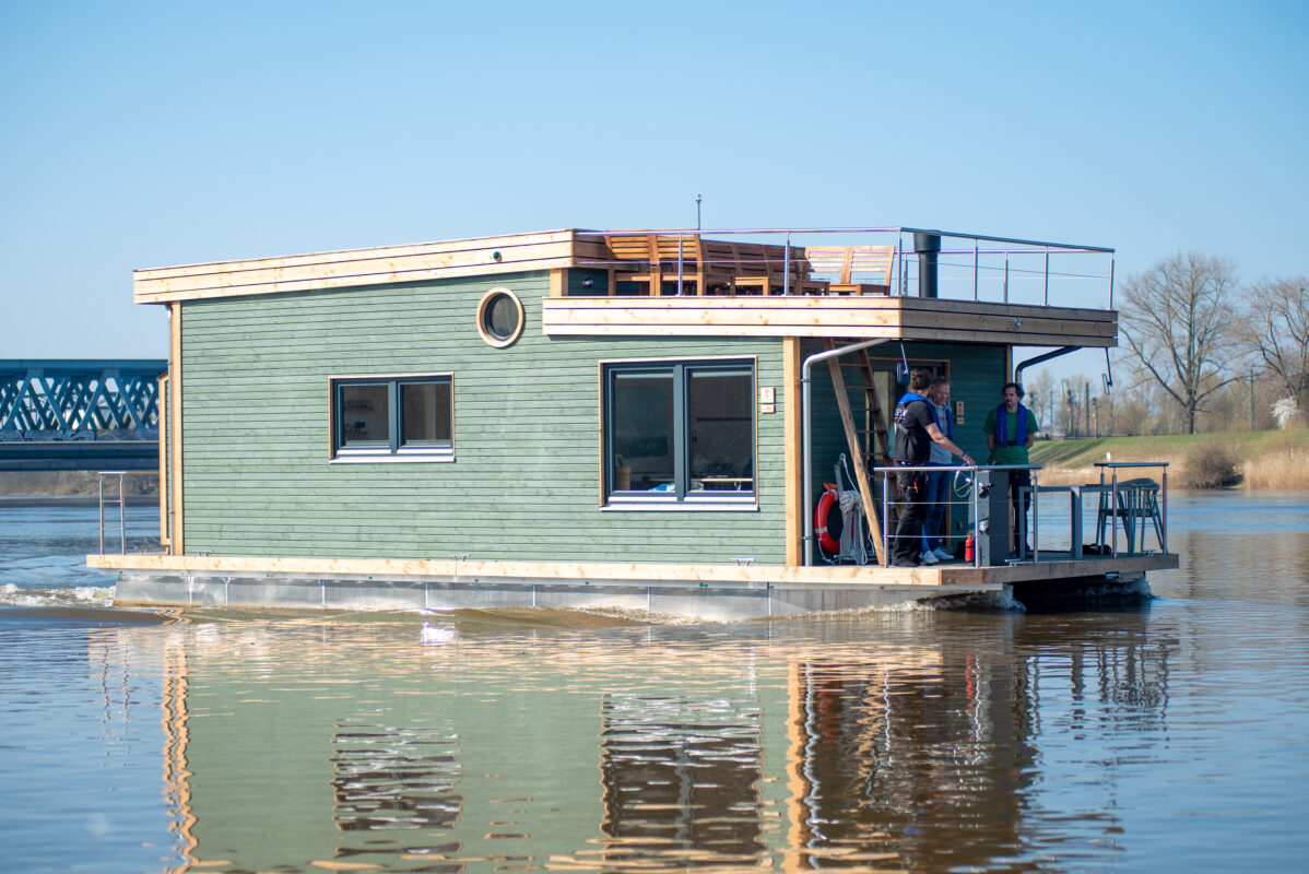 modernes grünes Hausboot, fahrend auf ruhigem Gewässer. Seitenansicht mit Dachterrasse und drei Personen am Steuerstand.
